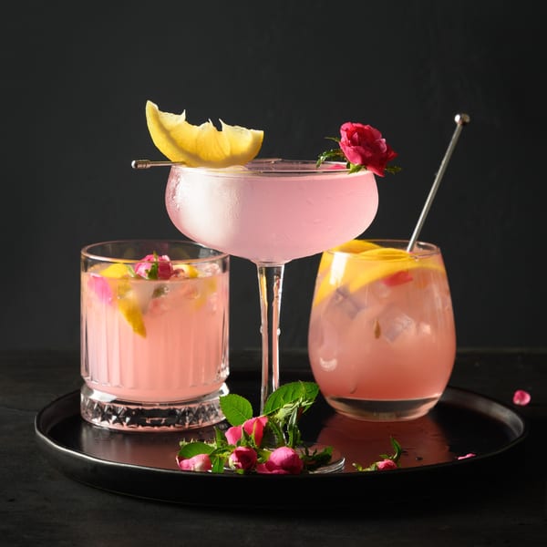 Three pink cocktails garnished with lemon slices and rosebuds on a black tray.
