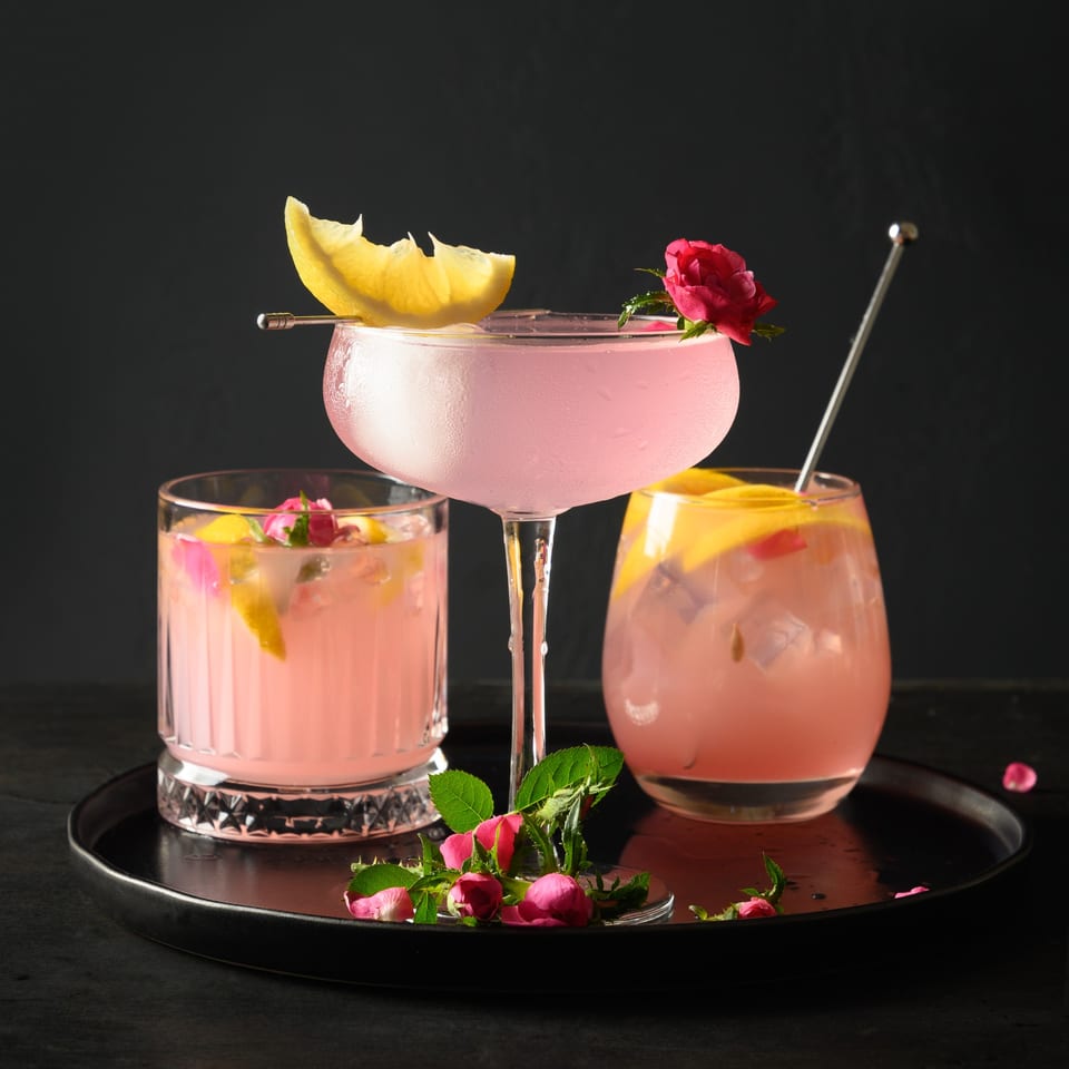 Three pink cocktails garnished with lemon slices and rosebuds on a black tray.